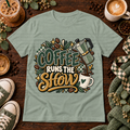 Sage green graphic T-shirt with a moka pot, coffee cup, coffee beans, and the phrase Coffee Runs the Show