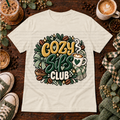 Cream graphic T-shirt with a coffee cup, scarf, coffee beans, bow, and the phrase Cozy Sips Club